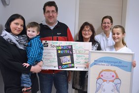 Spende Muko (Krankenhaus Barmherzige Brüder Regensburg) Auf dem Bild ist der kleine Dominik mit seine Eltern zu sehen. Seine Mama hält ihn im Arm. Der Vater hält den Scheck und drei andere Personen freuen sich mit ihnen.