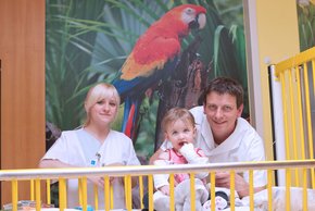 Kinderkrankenpflege (Krankenhaus Barmherzige Brüder Regensburg) Auf dem Bild sieht man rechts einen braunhaarigen Arzt, mittig ein Baby und links eine blonde Krankenschwester, die auf einem Bett knien. Im Hintergrund sieht man ein Foto eines Papageis.