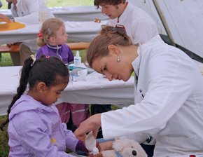 Teddyklinik (Krankenhaus Barmherzige Brüder Regensburg) Man sieht eine Frau im Doktorkittel, die einen Teddy untersucht. Ein kleines Mädchen sieht ihr interessiert zu.