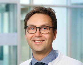 Badelt (Clemens Mayer) Herr Badelt trägt eine schicke Hornbrille, ein blau kariertes Hemd und passende blaue Krawatte. darüber einen Arztkittel. Er lächelt sehr freundlich