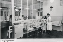 Säuglingszimmer (Orden der Blauen Schwestern der Heiligen Elisabeth) Man sieht eine schwarz weiß Aufnahme. Darauf sind mehrere kleine Betten abgebildet und eine Schwester plus Arzt mit Baby im Arm.